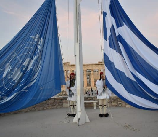 Δύο τσολιάδες στην Ακρόπολη ανυψώνουν τη σημαία της Ελλάδας και του ΟΗΕ, με φόντο τον Παρθενώνα, σε μια τελετουργική στιγμή που συμβολίζει τη διεθνή συνεργασία και την ελληνική πολιτιστική κληρονομιά.