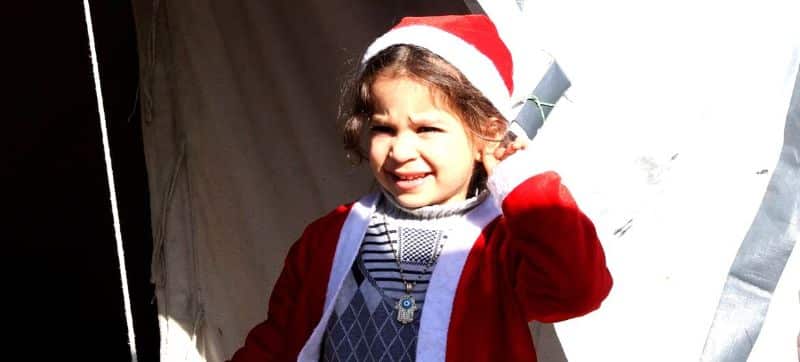 UN News | A young Palestinian girl wearing a Santa Claus outfit, stands in front of tents belonging to displaced families in northern Gaza City.