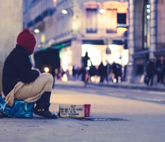 Une personne sans abri regardant vers des magasins est assise dans un boulevard haussmannien.