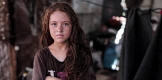 Portrait en gros plan d'une jeune fille au regard sérieux et direct fixé op l'objectif. Elle a de longs cheveux ondulés châtain-roux et de nombreuses taches de rousseur sur le visage. Elle se tient les bras croisés dans un espace intérieur sombre et encombré, sur un fond flou. Elle porte un t-shirt simple, marron foncé, avec un imprimé partiellement visible.