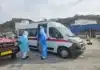 Wereldgezondheidsorganisatie: hantavirus-respons op besmettingen van cruiseschip Medische evacuatie in een haven in Kaapverdië op 6 mei 2026. Drie hulpverleners in volledige blauwe en witte beschermende pakken (PPE) staan bij de open schuifdeur van een witte ambulance. Eén hulpverlener houdt een brancard vast, terwijl een ander in de ambulance stapt. Links staat een rood voertuig met de tekst 'AMBULANCE' in spiegelbeeld op de motorkap. Op de achtergrond zijn een havenkade, vissersboten en gebouwen tegen een heuvel zichtbaar.