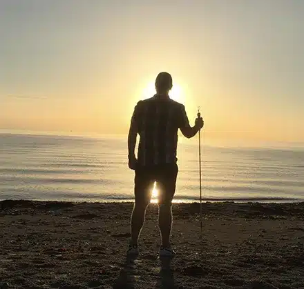 En man med vit käpp i en solnedgång på stranden. 